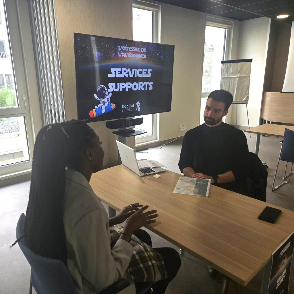 1 étudiant entrain de passer un entretien avec un ordinateur dans une salle avec un écran TV